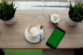 White cup of hot cappuccino on white saucer, black mobile phone, red velvet dessert and green grass in pots on wooden bar table Royalty Free Stock Photo