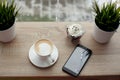 White cup of hot cappuccino on white saucer, black mobile phone, red velvet dessert and green grass in pots on wooden bar table Royalty Free Stock Photo