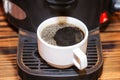 A white cup with prepared black coffee is in the coffee machine. Breakfasts and business meetings. Close-up