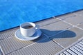 A white cup on a saucer with coffee stands at the edge of the pool. The cup casts a shadow