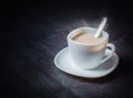 White cup on a saucer with hot cocoa. Morning breakfast. Dark wooden background.