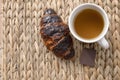 White cup of tea with a crispy croissant and a square of chocolate placed on a wicker mat Royalty Free Stock Photo