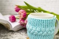 White cup of tea in a knitted blue case with an open book and with pink flowers tulips and sweets, with gingerbread on a Royalty Free Stock Photo