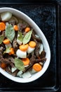 White dish with meat, carrots, onions and laurel on a black pan.