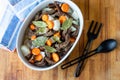 White dish with meat, carrots, onions and laurel on a wooden background. Cooking appliances spoon and fork. Top view.