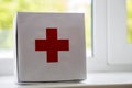 White first aid kit with red cross indoors on windowsill on blurred background. Healthcare concept