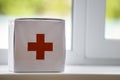 White first aid kit with red cross indoors on windowsill on blurred background. Healthcare concept