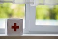 White first aid kit with red cross indoors on windowsill on blurred background. Healthcare concept