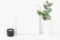 White frame mockup composition with branches of green silver dollar eucalyptus in ceramic vase black candle on table wall