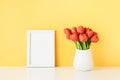 White frame mockup and red tulips in a white vase on a white table by the yellow wall Royalty Free Stock Photo