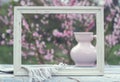 White frame, porcelain pink jug, cup and napkin on a table of white boards against the background of a flowering bush
