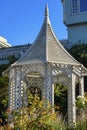 White gazebo with tall spire and top front yard trees and plants with urban or suburban background blue sky downtown