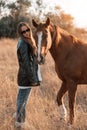 White girl in black leather jacket and glasses next to a horse and poses in the forest at sunset, like in a fairy tale Royalty Free Stock Photo