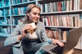 White girl near bookshelf in library. Student is eating sandwitch and using laptop.