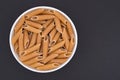 White glass bowl of raw brown penne rigate pasta. Isolated on a dark grey background. Royalty Free Stock Photo