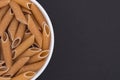 White glass bowl of raw brown penne rigate pasta. Isolated on a dark grey background. Royalty Free Stock Photo