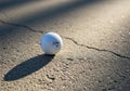 A white golf ball is resting on a textured, cracked pavement surface. The ball casts a shadow Royalty Free Stock Photo