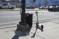 A white and green electric scooter next to a black metal trash can on the sidewalk downtown next to a tall black pole