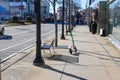 A white and green electric scooter on the sidewalk in front of a black metal bench surrounded by bare winter trees and buildings