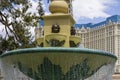 A white and green marble water fountain with flowing water surrounded by lush green trees and plants with people, blue sky