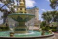 A white and green marble water fountain with flowing water surrounded by lush green trees and plants with people, blue sky