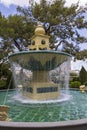 A white and green marble water fountain with flowing water surrounded by lush green trees and plants with people, blue sky