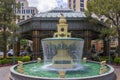 A white and green marble water fountain with flowing water surrounded by lush green trees and plants with people, blue sky