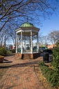 A white and green wooden gazebo on a red brick footpath surrounded by bare winter trees and lush green plants
