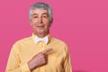 White haired mature man points aside at upper right corner, dresses yellow shirt with bow tie, isolated over pink background. Royalty Free Stock Photo