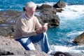 White-haired senior man  sitting on the rocks enjoying the sun in a winter day - using mobile phone and smiling. Relaxed elderly Royalty Free Stock Photo