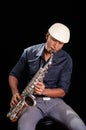 With white hat a black man is sitting and playing his saxophone, dark background, nice music Royalty Free Stock Photo