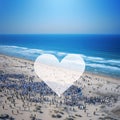 White heart against a background of hundreds of people gathered on the beach against the sea. Heart as a symbol of affection and