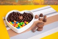 White heart-shaped bowl filled with brown and colorful chocolates, hazelnuts and almonds covered in chocolate