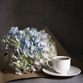 White hydrangea in a glass vase with a small cup of coffee on a book with a notebook on a gray craft background