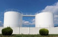 White industrial liquid storage tanks with a white fence in front. Blue sky with fluffy clouds on background