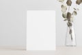 White invitation card mockup with an eucalyptus on a beige table. 5x7 ratio, similar to A6, A5