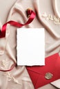 White invitation card mockup, red envelope and ribbon on beige silk. Flat lay, top view, copy space. Elegant wedding stationery Royalty Free Stock Photo