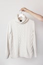 White knitted wool sweater on a white wooden hanger in female hands against the background of the light wall. Autumn and winter cl