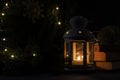 White lantern with a burning candle. Nooks, christmas tree branch and lights on background. Night dark picture. Christmas mystery.