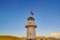 A white lighthouse stands against a blue sky. Over the lighthouse a red Turkish flag flies. Behind the lighthouse you can see the