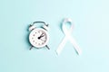 White lung cancer awareness ribbon and alarm clock on blue background Royalty Free Stock Photo