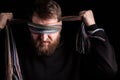 A white man with a beard blindfolds himself with a multi-colored scarf