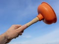 A white man`s hand raises an orange plunger towards the blue sky, holding it by the wooden handle