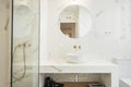 White marble bathroom with round mirror, gold faucets and shower