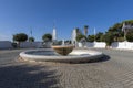 A white, marble fountain standing in a small circular square. Clear water gushes from the fountain