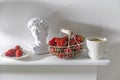 White metal basket with wooden handle with fresh strawberries, corrugated ceramic cup of tea, plaster Apollo head on a beige table