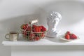 White metal basket with wooden handle with fresh strawberries, corrugated ceramic cup of tea, plaster Apollo head on a beige table
