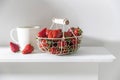 White metal basket with wooden handle with fresh strawberries and corrugated ceramic tea cup on beige table