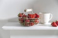 White metal basket with wooden handle with fresh strawberries and corrugated ceramic tea cup on beige table