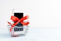 White mobile phone with red ribbon in shopping basket on white background. Gift, sale, e-commerce concept Royalty Free Stock Photo